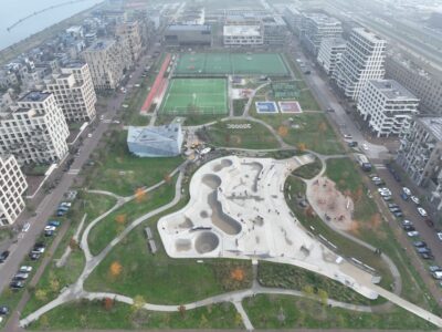 Luchtfoto van het recreatiepark op Zeeburgereiland in Amsterdam. Op de voorgrond een groot modern betonnen skatepark met bowls, ramps en plateaus waar mensen skaten, omringd door groen, paden en bomen. Op de achtergrond meerdere sportvelden (voetbalvelden met kunstgras en basketbalcourts), geflankeerd door moderne woonappartementen en straten met geparkeerde auto’s.